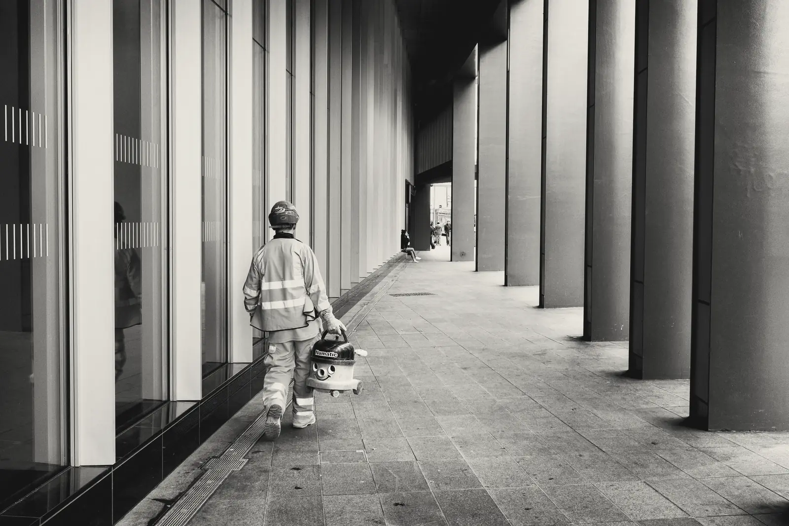 A construction worker in hi-vis walks along a colonnade of tall columns, carrying a vacuum cleaner with a smiling face.