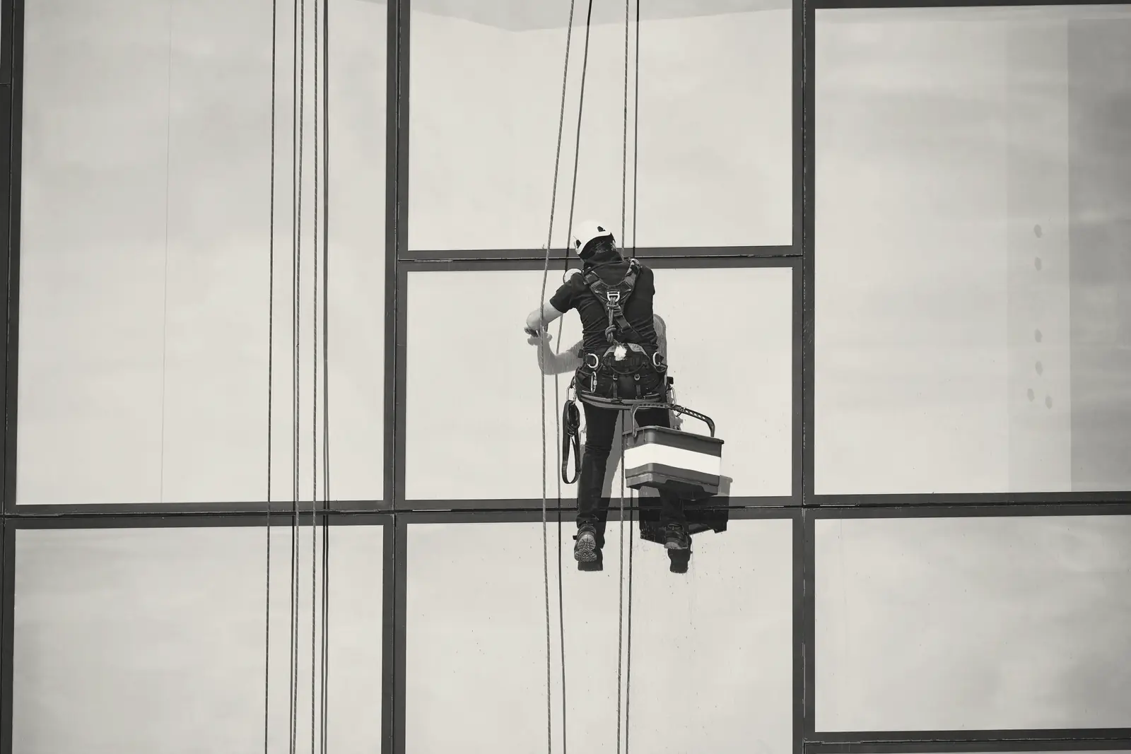 A rope-access window cleaner in a harness hangs against the glass facade of a tall building, bucket at his side.