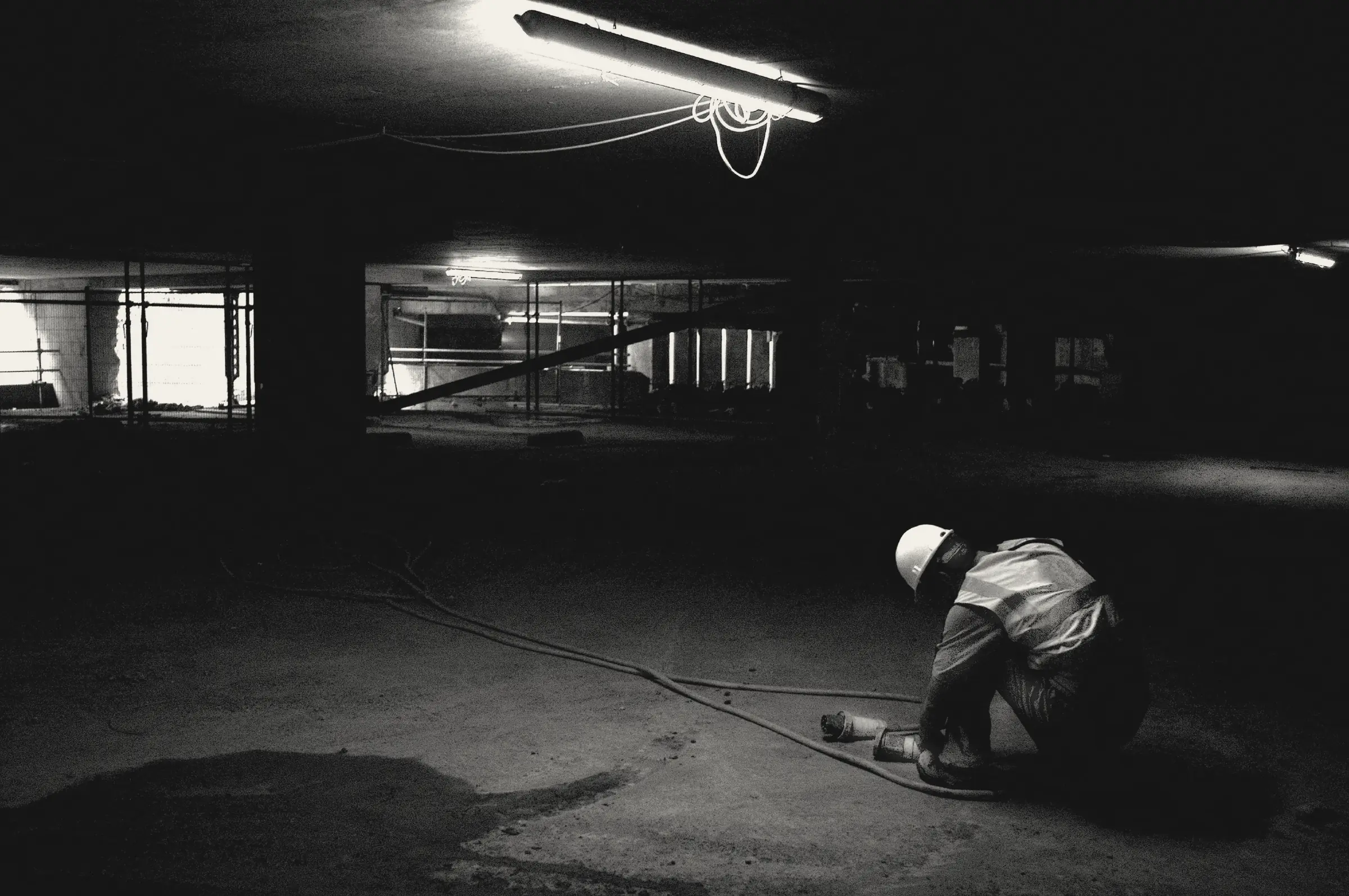 A lone construction worker in a hard hat and hi-vis vest crouches on the floor of a dark underground site, lit by a single strip light.