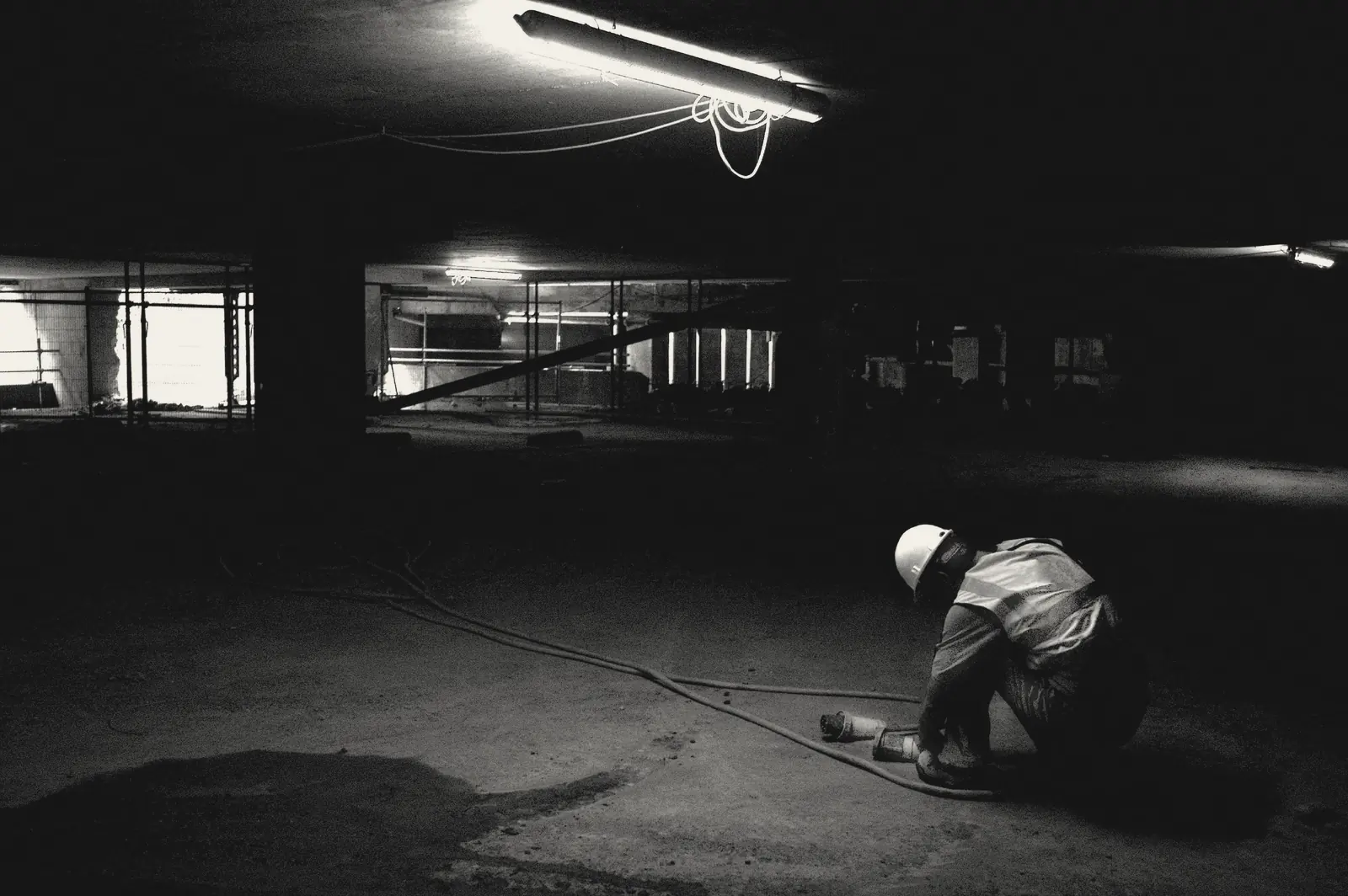 A lone construction worker in a hard hat and hi-vis vest crouches on the floor of a dark underground site, lit by a single strip light.